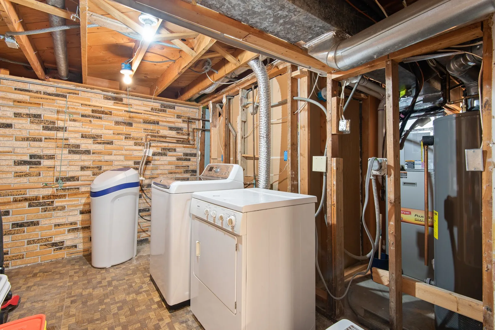 An unfinished basement utility area with a washer and dryer, water softener, exposed joists and ductwork, and a conventional tank water heater beside the furnace.
