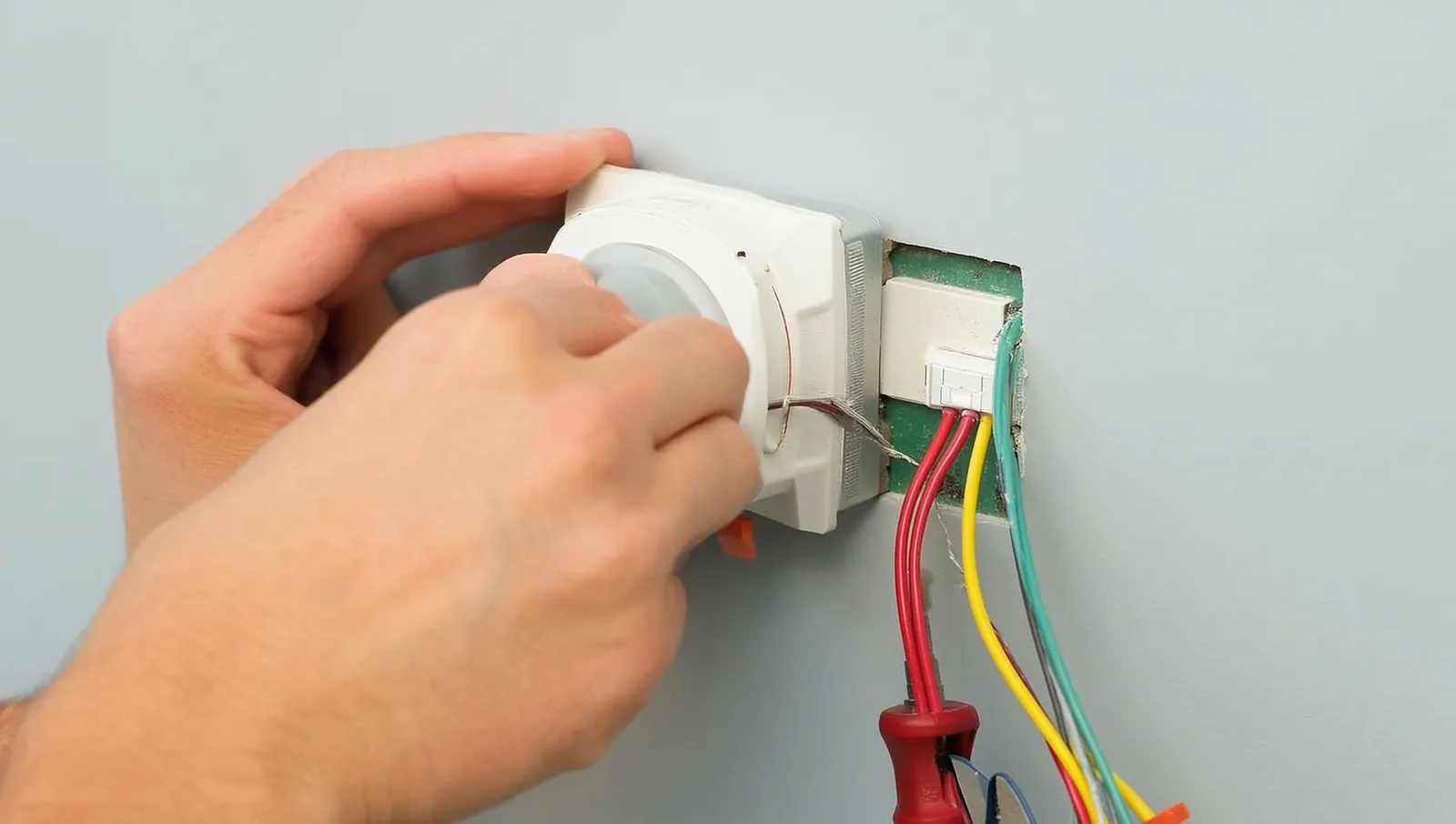 A person's hands wiring a smart thermostat base plate to the low-voltage wires coming out of the wall, with a screwdriver resting nearby.