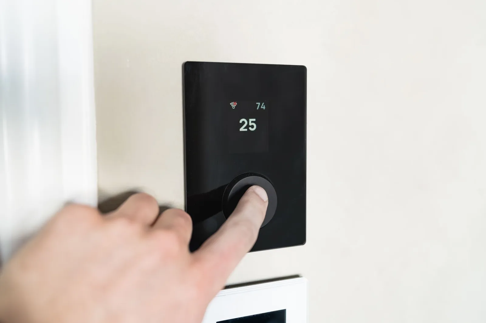 A hand pressing the dial on a sleek black wall-mounted smart thermostat showing a temperature reading.